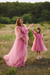 Fototapeta na wymiar Mother and her daughter in tulle pink dresses posing on nature at summer. Beautiful mummy holding hand of her little girl outdoor. Love. Family concept