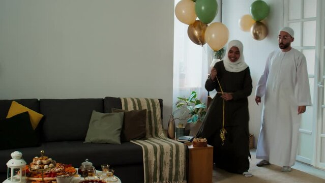 Young Muslim husband and wife in traditional Arabian clothes decorating living room with balloons while woman in hijab putting bunch between couch and served table