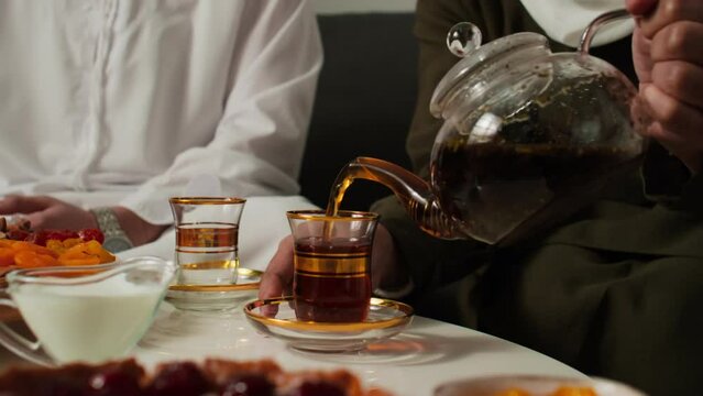 Young unrecognizable Muslim woman pouring hot black tea from teapot into glass while sitting by served table next to her husband wearing white kandura