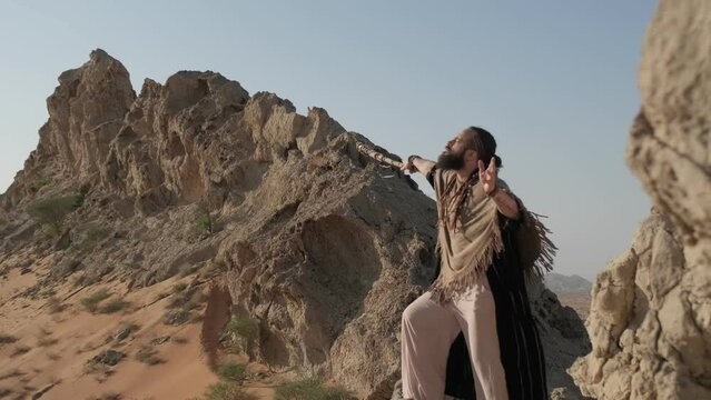 A man with a staff in his hand and the clothes of a shaman stands on a rock among the sands of the desert and dances