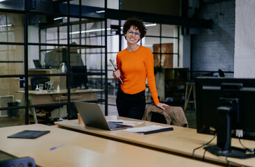 Fototapeta na wymiar Cheerful young woman holding tablet while standing by table with gadgets in office