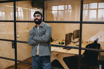 Confident bearded man standing near glass wall while folding arms in office
