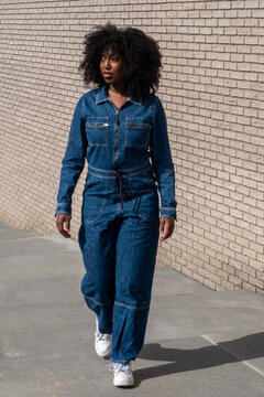 A stylish woman with natural afro hair strides confidently in a chic denim jumpsuit. The simplicity of the brick wall backdrop emphasizes her fashionable outfit and the effortless cool of her urban