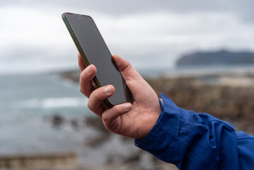 Close-up of a man's hand holding his mobile phone with a natural coastal landscape in the background out of focus on a cloudy day