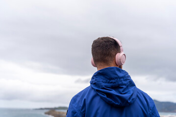 man on his back with pink headphones and a blue jacket enjoying the music while looking at the horizon and the cloudy sky in the background.
