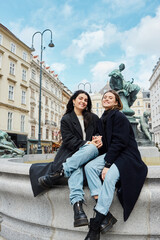 happy lesbian couple holding hands and sitting by a fountain statue with cityscape in Vienna