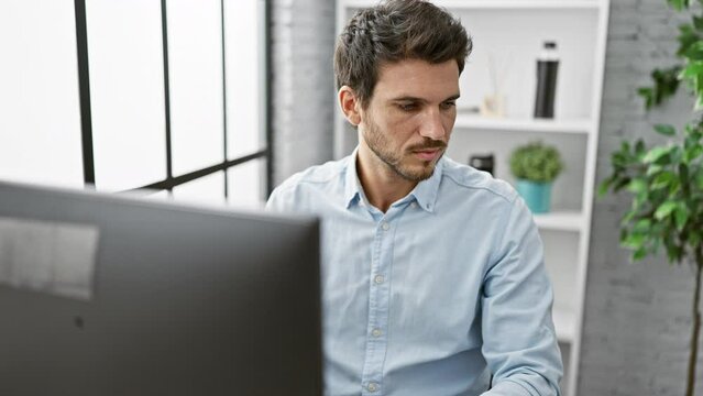 Focused hispanic man with beard works at computer in modern office setting, reflecting professional and urban lifestyle.