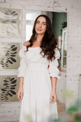 Women's day. Romantic attractive young woman 20-25 years with long curly hair in white dress is poses with crossed hands and looks at camera at modern stylish interior of home kitchen on background.