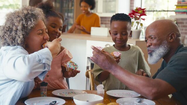 Grandparents with grandchildren indoors at home decorating cupcakes with icing and sprinkles together with parents in background - shot in slow motion