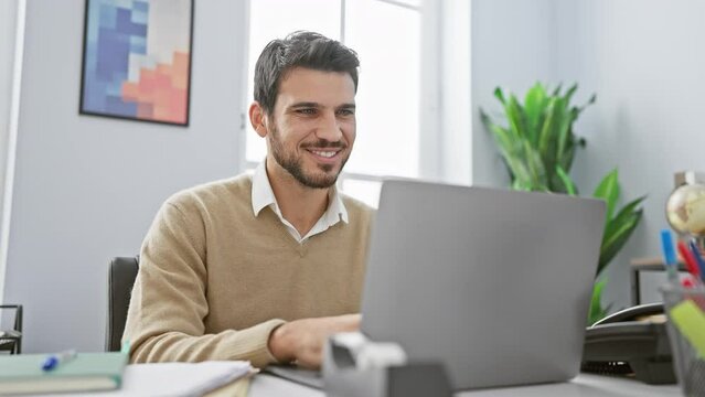 Smiling young hispanic man with beard working on laptop in modern office interior