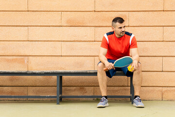 An adult male in his 30s and 40s is playing pickleball on an outdoor track at sunset.The young man is sitting on a bench waiting for the game to start.Pickle ball concept.Men playing pickleball.