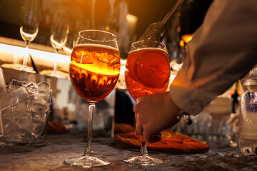 Bartender makes two glasses of cocktail Aperol spritz on bar counter, adds fresh orange slices....
