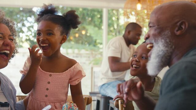 Grandparents with messy grandchildren indoors at home decorating cupcakes with icing and sprinkles together with parents in background - shot in slow motion