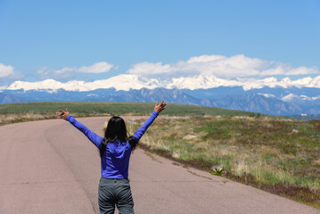 A woman in a blue shirt with arms outstretched toward the sky with the Rocky mountain range in the...
