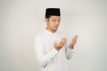 Asian muslim man is praying with hands up, wearing Arab costume in copy space on isolated background - obrazy, fototapety, plakaty