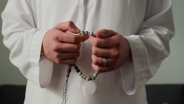 Hands of young unrecognizable Muslim man in white thobe moving his fingers over grey rosary beads while standing in front of camera during namaz prayerHands of young unrecognizable Muslim man in white