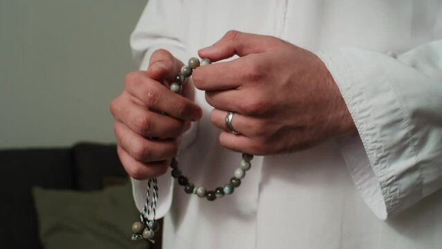 Hands of young Muslim man in white kandora fingering grey beads of rosary in front of himself while standing in living room against couch and performing namaz prayer