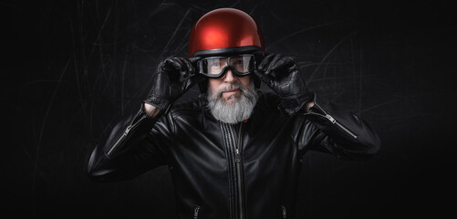 Portrait d'un motard homme avec un casque rouge et une veste en cuir et des lunettes de protection et une barbe grise vintage - 769570148