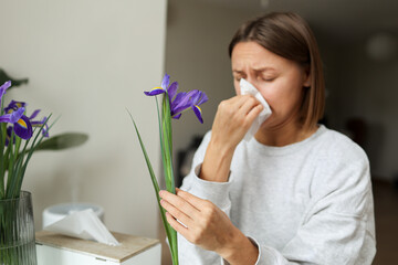 Allergic young woman holds iris flower, covers nose with paper tissue has runny nose, sneezes from...