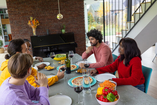 An engaging snapshot of a diverse group of friends engrossed in conversation over a delicious lunch in a stylish home setting. The table is rich with Italian-inspired dishes, and the open room is