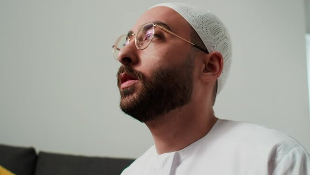 Tilt up and down of Muslim man in traditional white kandora and skullcap standing on his knees in fornt of camera in home environment and moving fingers over rosary beads during prayer