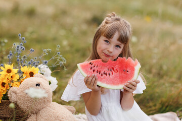 Adorable little child girl bites big slice of watermelon at picnic with teddy bear, wild flowers outdoors on sunny day. Healthy fruit snack for children. Summer holidays concept and happy childhood.