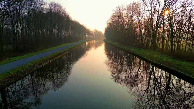 This captivating drone footage captures the serene beauty of a tranquil canal flanked by rows of bare trees, reflecting the soft glow of the rising or setting sun. The calm waters mirror the pastel