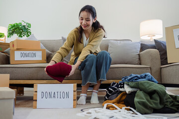 Asian young woman packing clothes at home, putting on stuff into donate box with second hand clothes. Help poor people. Donation concept - obrazy, fototapety, plakaty