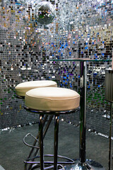 Two white bar stools and a glass tabletop on a silver shiny background of small foil squares. There is a disco ball hanging above the bar
