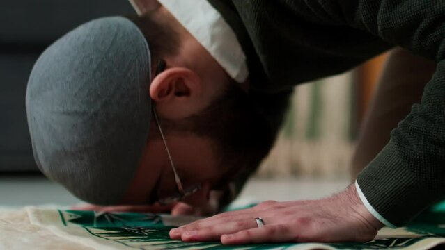 Young religious man in grey skullcap and pullover standing on knees in front of camera in home environment and touching rug by his head during silent prayer