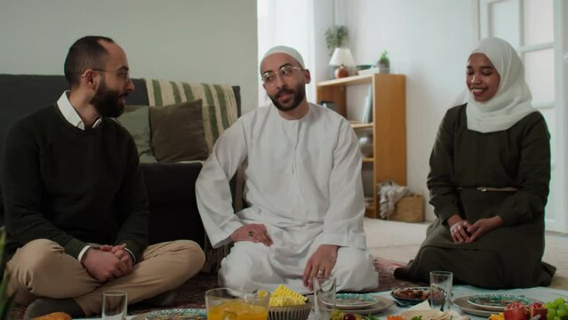 Young Muslim man in white kandora and skullcap talking to his twin brother in casualwear while woman in hijab sitting next to her husband during family dinner