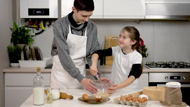Happy and smiling girl whisking egg in bowl with metal wire whisk. Sister helps brother in kitchen. Teenage children cooking process cookies and pastry. Close knit family and happy childhood