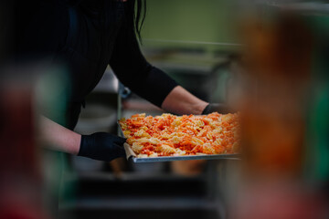 A person in black gloves is handling a tray of orange freeze-dried carrot slices in a blurred kitchen setting.