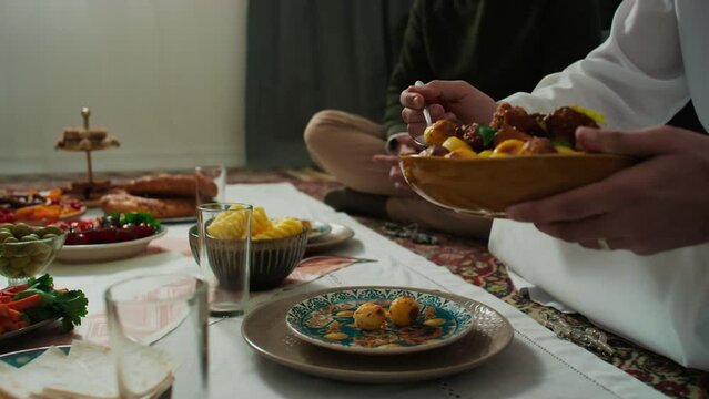 Hands of young unrecognizable hospitable man in Muslim clothes holding plate with baked homemade meal and putting potatoes to his guest sitting on the floor in front of tablecloth with food