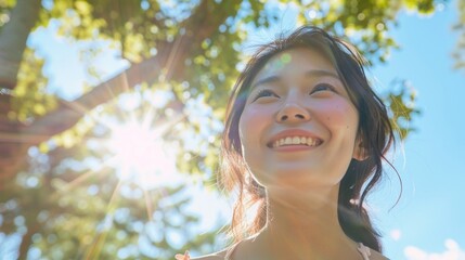 A young woman with a radiant smile looking up at the sun through the dappled light of a tree canopy on a clear day.