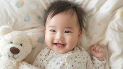 A joyful baby with a big smile lying on a patterned blanket next to a white teddy bear.