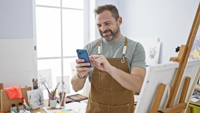 A handsome middle-aged man with grey hair wearing an apron using a smartphone in a bright art studio