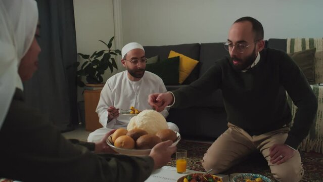 Young man taking two flatbreads being passed by hospitable Muslim woman in hijab and long dress over tablecloth with homemade snacks and drinks during family dinner