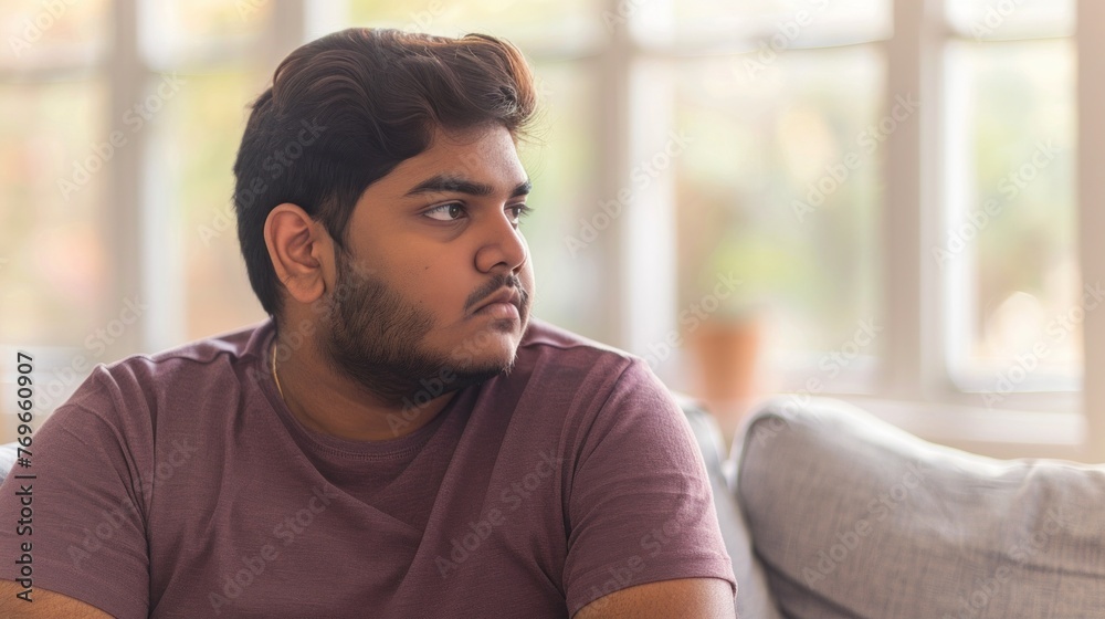 Poster Young man with beard sitting on couch looking away from camera in room with large windows.