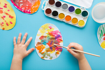 Baby boy holding paintbrush and painting colorful egg on paper with watercolor on pastel blue table...