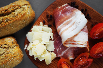 Wooden board with slices of bacon, parmesan, cherry tomatoes, black peppercorns next to bread and knife on gray background