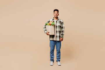 Full body smiling happy fun young man wear grey shirt hold paper bag for takeaway mock up with food products look camera isolated on plain beige background. Delivery service from shop or restaurant.