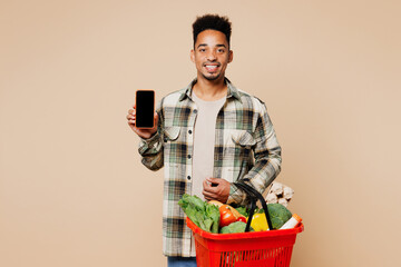 Young man wear grey shirt hold red basket bag with food products use blank screen workspace area mobile cell phone isolated on plain light beige background. Delivery service from shop or restaurant.