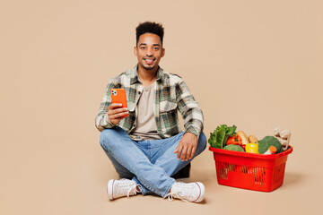Full body smiling happy young man wears grey shirt sit near red basket bag with food products hold use mobile cell phone isolated on plain beige background. Delivery service from shop or restaurant.