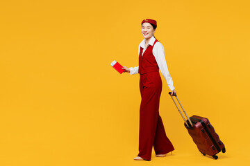 Full body young stewardess flight attendant woman of Asian ethnicity wear red vest shirt hat hold bag passport ticket travel abroad isolated on plain yellow background studio. Air flight trip concept.