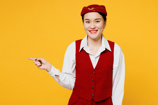Young stewardess flight attendant woman of Asian ethnicity she wears red vest shirt hat point index finger aside on area isolated on plain yellow background studio portrait. Air flight trip concept.