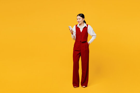 Full body smiling fun young lawyer employee business woman of Asian ethnicity wear formal red vest shirt work at office hold use mobile cell phone isolated on plain yellow background studio portrait