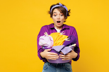 Young surprised shocked scared amazed woman wear purple shirt do housework tidy up hold look at basin with laundry clothes isolated on plain yellow background studio portrait. Housekeeping concept.