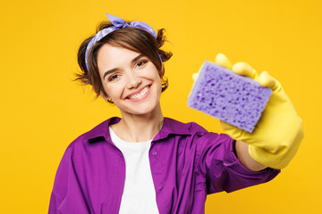Young smiling happy woman she wear purple shirt rubber gloves do housework tidy up hold in hand sponge stretch hand to camera isolated on plain yellow background studio portrait. Housekeeping concept.
