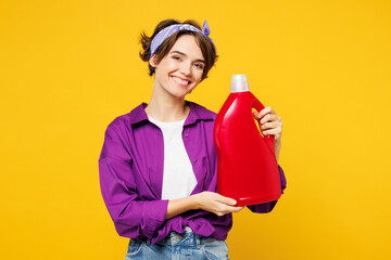 Young smiling fun woman wear purple shirt casual clothes do housework tidy up hold in hand fabric softener bottle look camera isolated on plain yellow background studio portrait. Housekeeping concept.